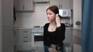 Genç kız zarif mutfakta birden fazla iş yapıyor, sesli mesajlar kaydediyor, modern yaşam tarzını ve verimliliğini cam masa dikey videosunda gösteriyor.