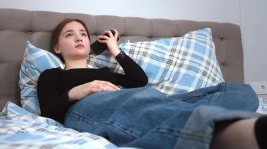 Rahat genç bir kız rahat bir şekilde yumuşak bir yatakta dinleniyor, mavi kot pantolon ve siyah gömlek giyiyor, akıllı telefonuyla sakin bir yatak odasında ekose yastıklarla iletişim kuruyor.