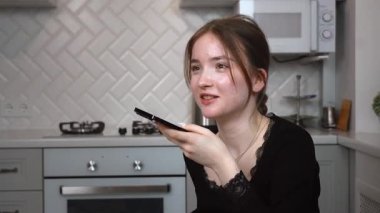 Genç kız zarif mutfakta birden fazla iş yapıyor, sesli mesajlar kaydediyor, modern yaşam tarzını ve verimliliğini cam masa dikey videosunda gösteriyor.