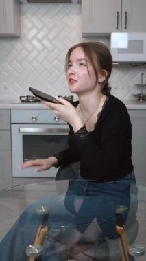 Genç kız zarif mutfakta birden fazla iş yapıyor, sesli mesajlar kaydediyor, modern yaşam tarzını ve verimliliğini cam masa dikey videosunda gösteriyor.