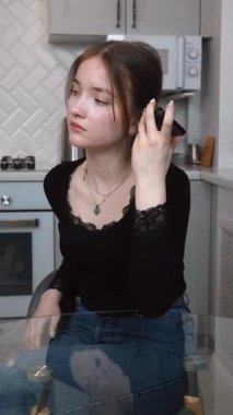 Genç bir kadın, muhtemelen bir vlogger ya da etkilici, modern bir mutfaktaki akıllı telefonuna sesli mesaj kaydediyor, ya da ses içeriği dikey video yaratıyor.