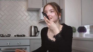 Genç bir kadın, muhtemelen bir vlogger veya etkilici, modern bir mutfaktaki akıllı telefonuna sesli mesaj kaydediyor, ya da ses içeriği yaratıyor.