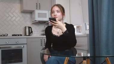 Genç kız akıllı telefonu yüze yakın tutuyor, sesli mesajlar kaydediyor ya da elleriyle konuşmuyor. Akıllı telefondaki sesli asistanı olan kadın rahat beyaz mutfak mobil iletişim uygulamasında