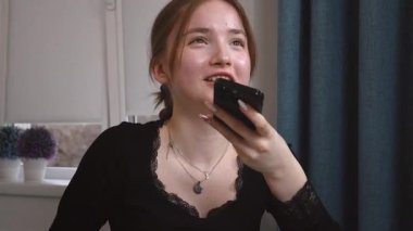 Genç kız akıllı telefonu yüze yakın tutuyor, sesli mesajlar kaydediyor ya da elleriyle konuşmuyor. Akıllı telefondaki sesli asistanı olan kadın rahat beyaz mutfak mobil iletişim uygulamasında