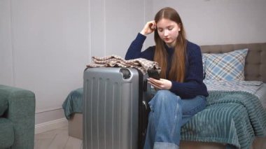 Düşünceli genç bir kadın, bir bavulun yanında yatarken akıllı telefonunu kontrol eder. Yolculuk için hazırlanmış gibi görünür. Seyahat detaylarını gözden geçirir ya da biriyle iletişim kurar.