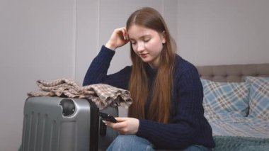 Düşünceli genç bir kadın, bir bavulun yanında yatarken akıllı telefonunu kontrol eder. Yolculuk için hazırlanmış gibi görünür. Seyahat detaylarını gözden geçirir ya da biriyle iletişim kurar.