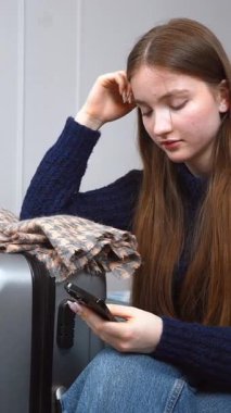 Düşünceli genç bir kadın, bir bavulun yanında yatarken akıllı telefonunu kontrol eder. Yolculuk için hazırlanmış gibi görünür. Seyahat detaylarını gözden geçirir ya da dikey görüntülerle iletişim kurar.