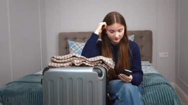 Düşünceli genç bir kadın, bir bavulun yanında yatarken akıllı telefonunu kontrol eder. Yolculuk için hazırlanmış gibi görünür. Seyahat detaylarını gözden geçirir ya da biriyle iletişim kurar.
