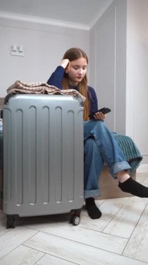 Düşünceli genç bir kadın, bir bavulun yanında yatarken akıllı telefonunu kontrol eder. Yolculuk için hazırlanmış gibi görünür. Seyahat detaylarını gözden geçirir ya da dikey görüntülerle iletişim kurar.