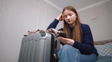 Genç kız, gündelik giysiler içinde otel odasında yatakta duruyor, bavulunu topluyor, uzun yolculuktan veya tatilden önce endişeli bir ifadeyle akıllı telefonu kontrol ediyor.