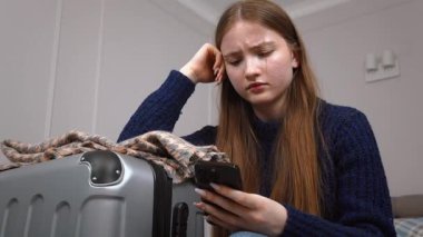 Genç kız, gündelik giysiler içinde otel odasında yatakta duruyor, bavulunu topluyor, uzun yolculuktan veya tatilden önce endişeli bir ifadeyle akıllı telefonu kontrol ediyor.