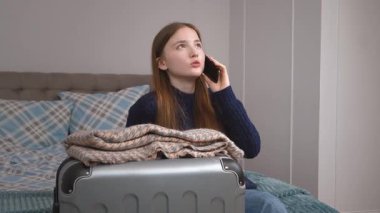 Uzun saçlı, günlük kıyafetlerle otel odasında oturan ve yanında cep telefonuyla konuşan genç bir kız, seyahat etmeye hazır bir bavul.