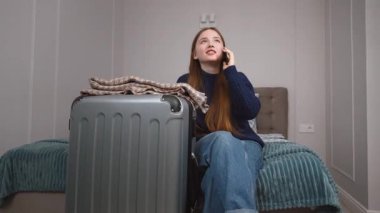 Uzun saçlı, günlük kıyafetlerle otel odasında oturan ve yanında cep telefonuyla konuşan genç bir kız, seyahat etmeye hazır bir bavul.