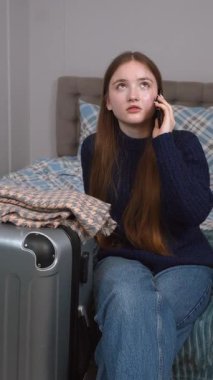 Uzun saçlı, günlük kıyafetleriyle otel odasında oturan ve yanında cep telefonuyla konuşan genç bir kız bavulu, dikey olarak seyahat etmeye hazır.