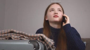 Uzun saçlı, günlük kıyafetlerle otel odasında oturan ve yanında cep telefonuyla konuşan genç bir kız, seyahat etmeye hazır bir bavul.