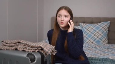 Akıllı telefonu ve bavuluyla otel odasında bavulunun yanında oturan genç bir gezgin kız telefonda turizm, seyahat, mutlu yaşam kavramı hakkında konuşuyor.