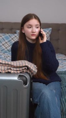 Uzun saçlı, günlük kıyafetleriyle otel odasında oturan ve yanında cep telefonuyla konuşan genç bir kız bavulu, dikey olarak seyahat etmeye hazır.