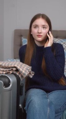 Akıllı telefonu ve bavuluyla otel odasında bavulunun yanında oturan genç bir gezgin kız telefonda turizm, seyahat ve mutlu bir hayat dikey videosu hakkında konuşuyor.