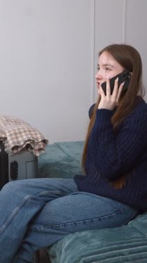 Akıllı telefonu ve bavuluyla otel odasında bavulunun yanında oturan genç bir gezgin kız telefonda turizm, seyahat ve mutlu bir hayat dikey videosu hakkında konuşuyor.