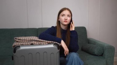 Akıllı telefonu ve bavuluyla otel odasında bavulunun yanında oturan genç bir gezgin kız telefonda turizm, seyahat, mutlu yaşam kavramı hakkında konuşuyor.