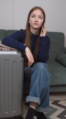 Akıllı telefonu ve bavuluyla otel odasında bavulunun yanında oturan genç bir gezgin kız telefonda turizm, seyahat ve mutlu bir hayat dikey videosu hakkında konuşuyor.