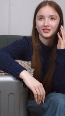 Akıllı telefonu ve bavuluyla otel odasında bavulunun yanında oturan genç bir gezgin kız telefonda turizm, seyahat ve mutlu bir hayat dikey videosu hakkında konuşuyor.