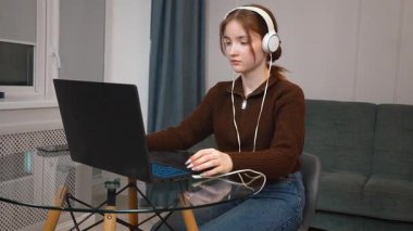 Evde kulaklık takıp bilgisayar oyunu oynayarak ya da iş yeri ya da sınav hazırlığı için dizüstü bilgisayar kullanarak uzaktan konsantre olan genç kız öğrenci.