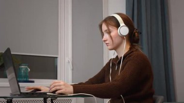 Evde kulaklık takıp bilgisayar oyunu oynayarak ya da iş yeri ya da sınav hazırlığı için dizüstü bilgisayar kullanarak uzaktan konsantre olan genç kız öğrenci.