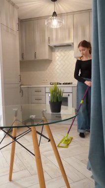 Genç bir kız mutfak zeminini paspasla siliyor. Modern yaşam alanlarını düzenli bir şekilde temizlik tekniğiyle temizliyor. Ebeveynlere temizlik konusunda yardım.