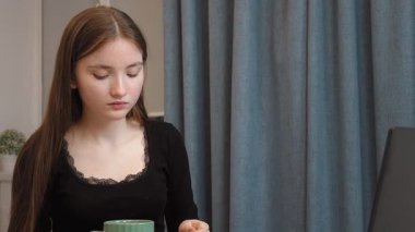 Genç kız laptopta çevrimiçi içeriği incelerken tatlı tatlının tadını çıkarıyor, dinlenme molası sırasında evde rahat bir şekilde oturuyor.