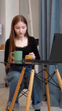 Modern mutfak ortamında, cam masada rahat bir şekilde oturan rahat ofis molası sırasında dizüstü bilgisayar başında çay ve pastanın keyfini çıkaran,