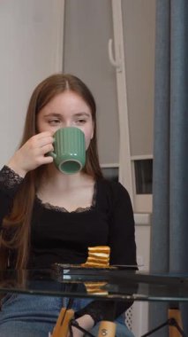 Genç bir kız bilgisayardaki çevrimiçi içeriği incelerken tatlı tatlının tadını çıkarıyor. Ev mutfağında rahatça oturup dinleniyor.