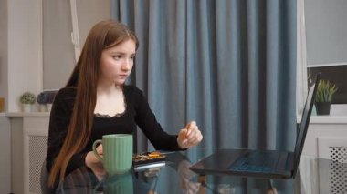 Genç kız laptopta çevrimiçi içeriği incelerken tatlı tatlının tadını çıkarıyor, dinlenme molası sırasında evde rahat bir şekilde oturuyor.