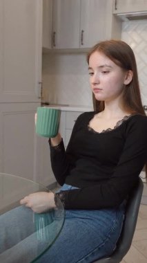 Uzun kahverengi saçlı genç kız modern mutfaktaki seramik bardaktan ılık bir içecek içiyor. Sabahın yumuşak, huzurlu atmosferinde ev yapımı dikey video var.