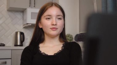 Genç kız elinde bir bardak sıcak içecekle modern mutfakta oturuyor ve bilgisayarla görüntülü konuşma yapıyor. Sorunları çözer, çalışır ve sadece akranlarıyla sohbet eder. dikey video