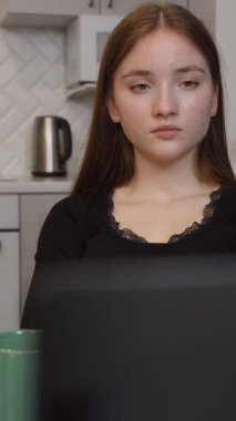 Genç kız elinde bir bardak sıcak içecekle modern mutfakta oturuyor ve bilgisayarla görüntülü konuşma yapıyor. Sorunları çözer, çalışır ve sadece akranlarıyla sohbet eder. dikey video