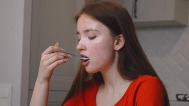 Genç kız, mutfaktaki cam masada oturuyor, turuncu tişört giyiyor ve yoğurt yiyor. Zevkle ve sakin bir ifadeyle, sağlıklı bir yaşam tarzını ve sağlıklı beslenme tarzını simgeliyor..