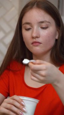 Genç kız, mutfaktaki cam masada oturuyor, turuncu tişört giyiyor ve zevk ve huzurlu bir ifadeyle yoğurt yiyor, sağlıklı bir yaşam tarzını ve sağlıklı beslenmesini kişileştiriyor. dikey video