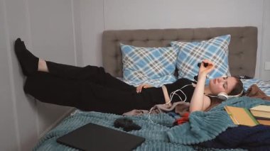 Kulaklıklı yorgun genç kız öğrenci ayakları duvara yaslanmış, kitaplar, giysiler ve dizüstü bilgisayarlarla çevrili, müzik dinliyor ve dersten sonra dinleniyor.