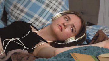 Genç kız öğrenci yatakta dağınık kulaklıklarla ve kitaplarla çevrili, yoğun çalışmadan sonra sakin müziğin tadını çıkarıyor.