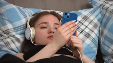 Kulaklıklı güzel bir genç kız, en sevdiği melodilere dalmış, gözleri kapalı yatakta yatarken akıllı telefondan müzik dinliyor.