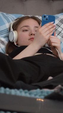 Akıllı telefondan müzik dinleyen kulaklıklı güzel bir genç kız gözleri kapalı, en sevdiği videoya dalmış bir şekilde yatakta uzanıyor.