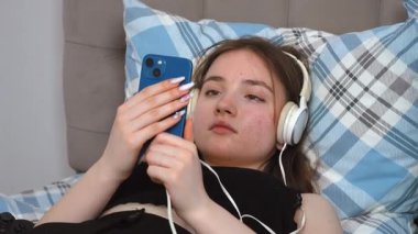 Kulaklıklı güzel bir genç kız, en sevdiği melodilere dalmış, gözleri kapalı yatakta yatarken akıllı telefondan müzik dinliyor.