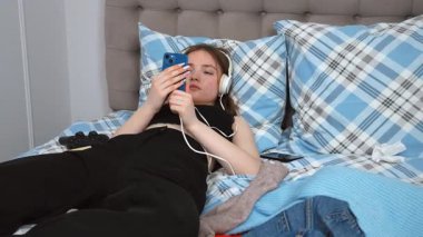 Kulaklıklı güzel bir genç kız, en sevdiği melodilere dalmış, gözleri kapalı yatakta yatarken akıllı telefondan müzik dinliyor.