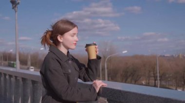 İçkiler ve insanların konsepti. Mutlu genç kadın ya da genç kız. Şehrin arka planında uzun saçlı, şık bir kızla kağıt bardaktan kahve içiyor.