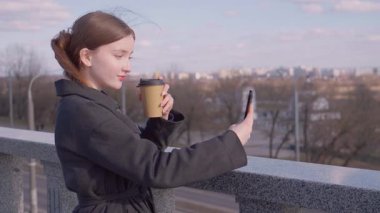 Genç bir kız akıllı telefon kamerasıyla şehir manzarasını filme çekerken paket kahvenin tadını çıkarıyor ve huzurlu bir görsel hikaye anlatma anında şehir balkonunun korkuluklarında dinleniyor.