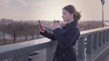 Genç bir kız şehirde dolaşırken, kahve içerken ve akıllı telefondan selfie çekerken resimli bir şehir manzarası ve nehir manzarasına bakan bir köprüde duruyor.