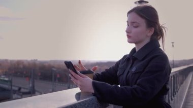 Sıradan giyinmiş genç bir kadın kahve eşliğinde rahatlıyor, yaya köprüsünde dikilirken akıllı telefona bakıyor, altın gün batımında şehir manzarasına bakıyor.
