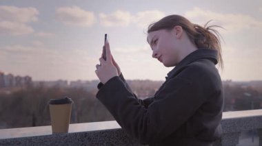 Kentsel yaşam tarzı kavramı, şehir manzarasına tepeden bakan bir balkonda kahve içerken fotoğraf çeken ya da telefonuyla video kaydeden genç bir kız tarafından örneklendirilir.