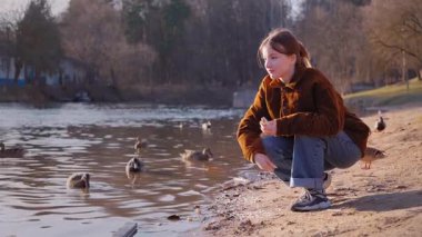 Gündelik giysiler giymiş genç bir kız su kenarında huzurlu bir anın tadını çıkarıyor, gölde yüzen ördekleri besliyor ve çömeliyor, doğanın barışçıl güzelliği ile çevrili.